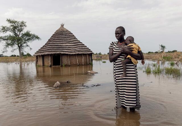 En kvinna håller sitt barn i famnen medans hon står framför sitt hus som är översvämmat i Sydsudan.