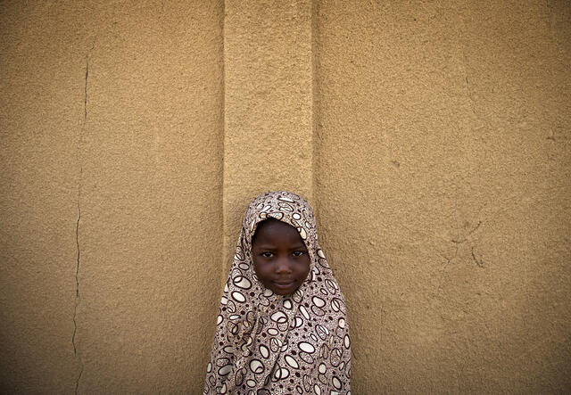 En flicka tittar in i kameran i Mali, hon står vid en husvägg.